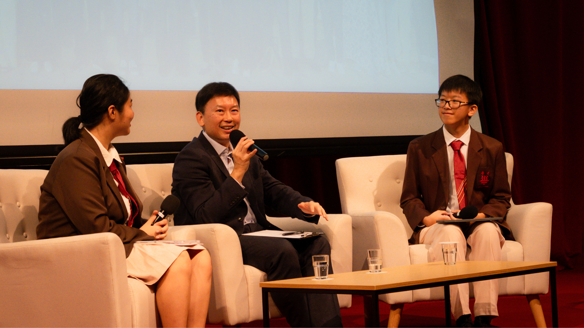Minister for National Development Mr. Chee Hong Tat speaking into a microphone while seated on stage for a dialogue session alongside two students in school uniform.
