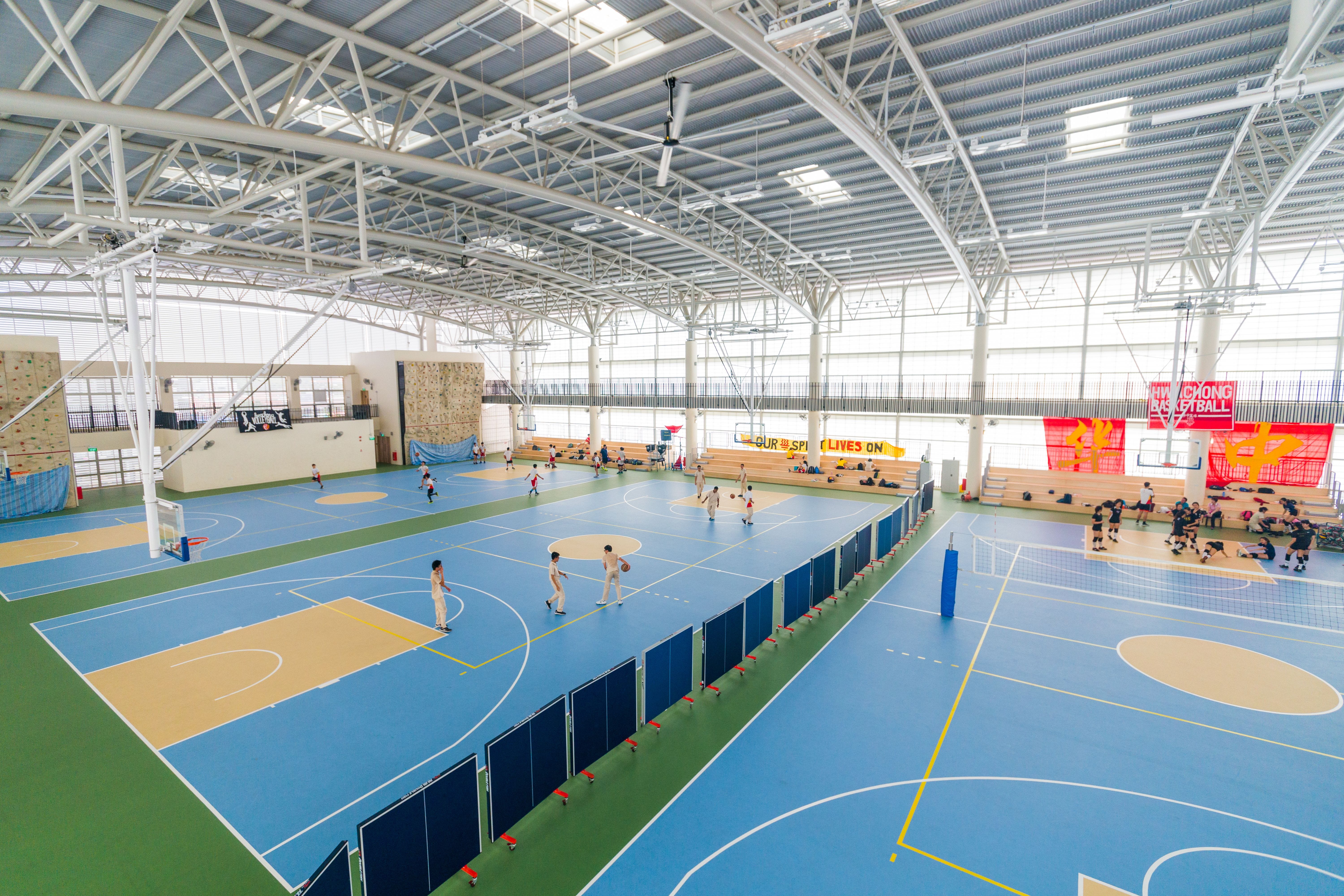 Large indoor sports hall with multiple blue basketball courts, players in action, a climbing wall, and bleachers along the side.
