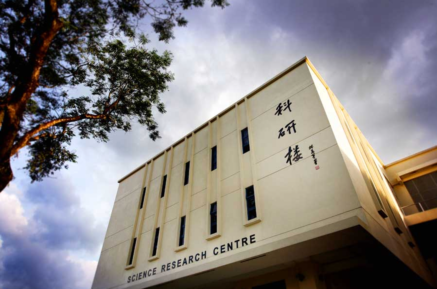 A cream-colored Science Research Centre building with Chinese characters, viewed from below against a cloudy sky with a tree.