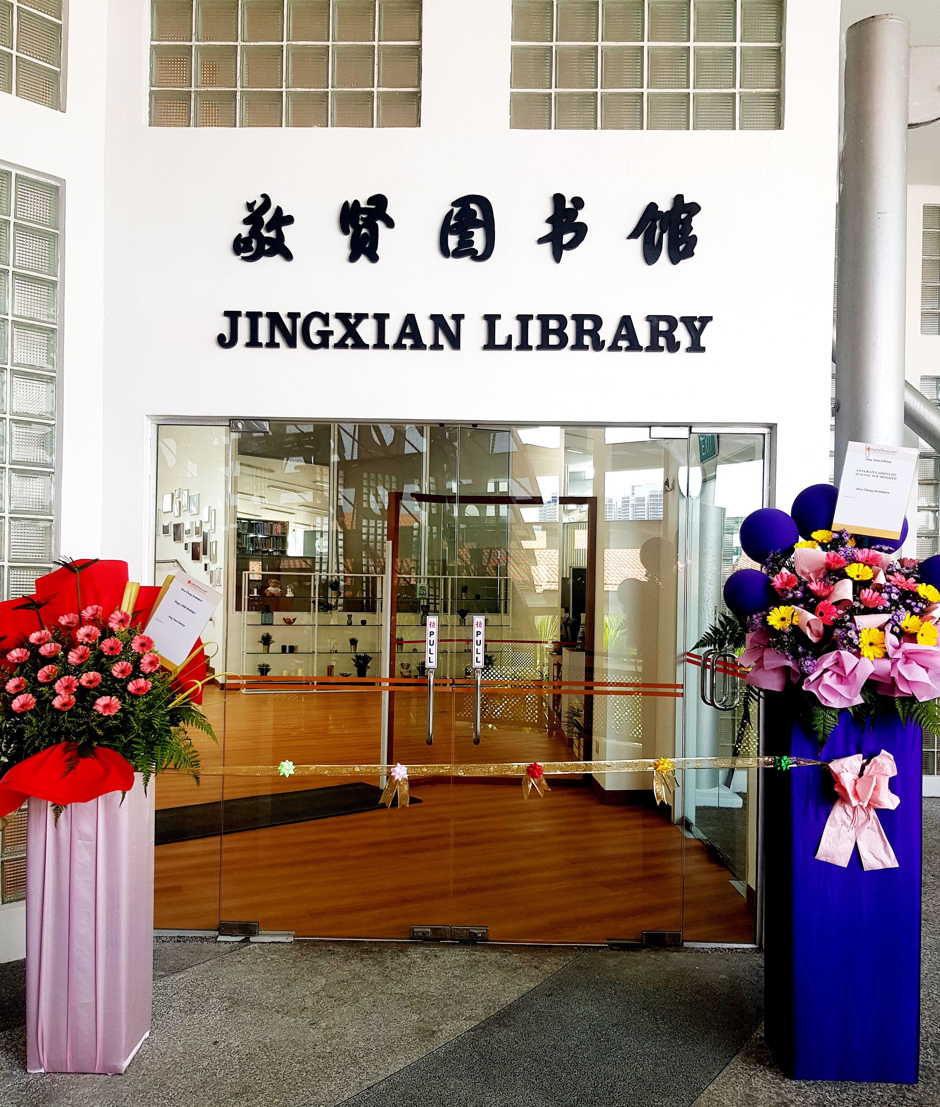 Entrance of Jingxian Library with Chinese characters above glass doors, flanked by two flower stands and purple balloons.