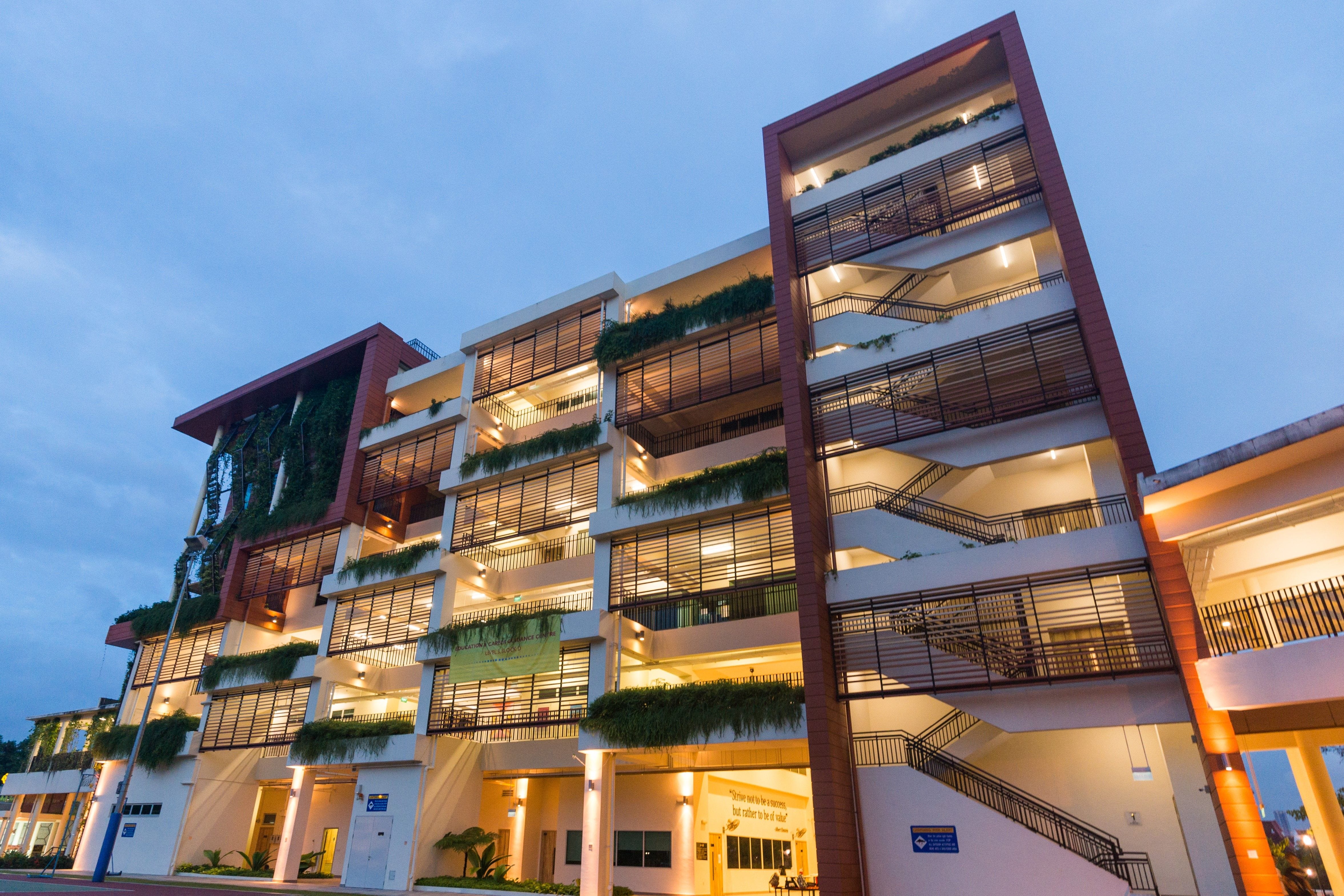 Modern multi-story building with green plants on balconies, warm interior lights, and a blue dusk sky.