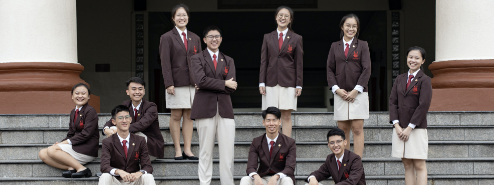 Nine students in matching dark red blazers and red ties pose on outdoor stone steps, smiling at the camera.