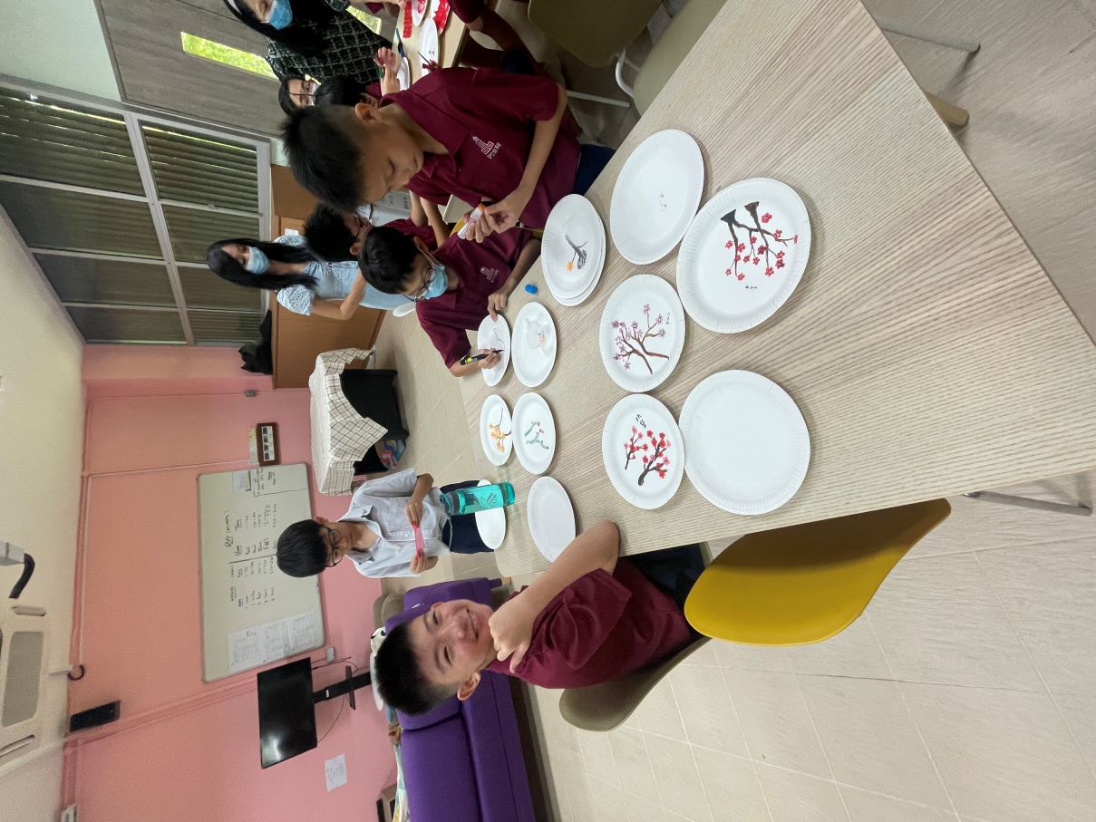 Children in a classroom decorate paper plates with blossom branch designs while teachers supervise