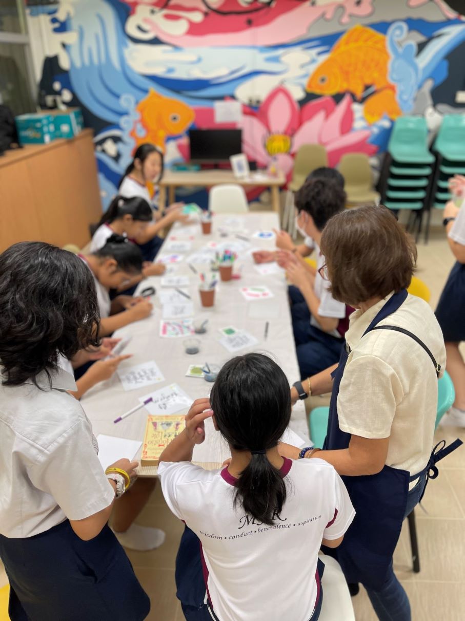 Students and an instructor gathered around a long table, participating in a hands-on art and craft activity in a colourful classroom