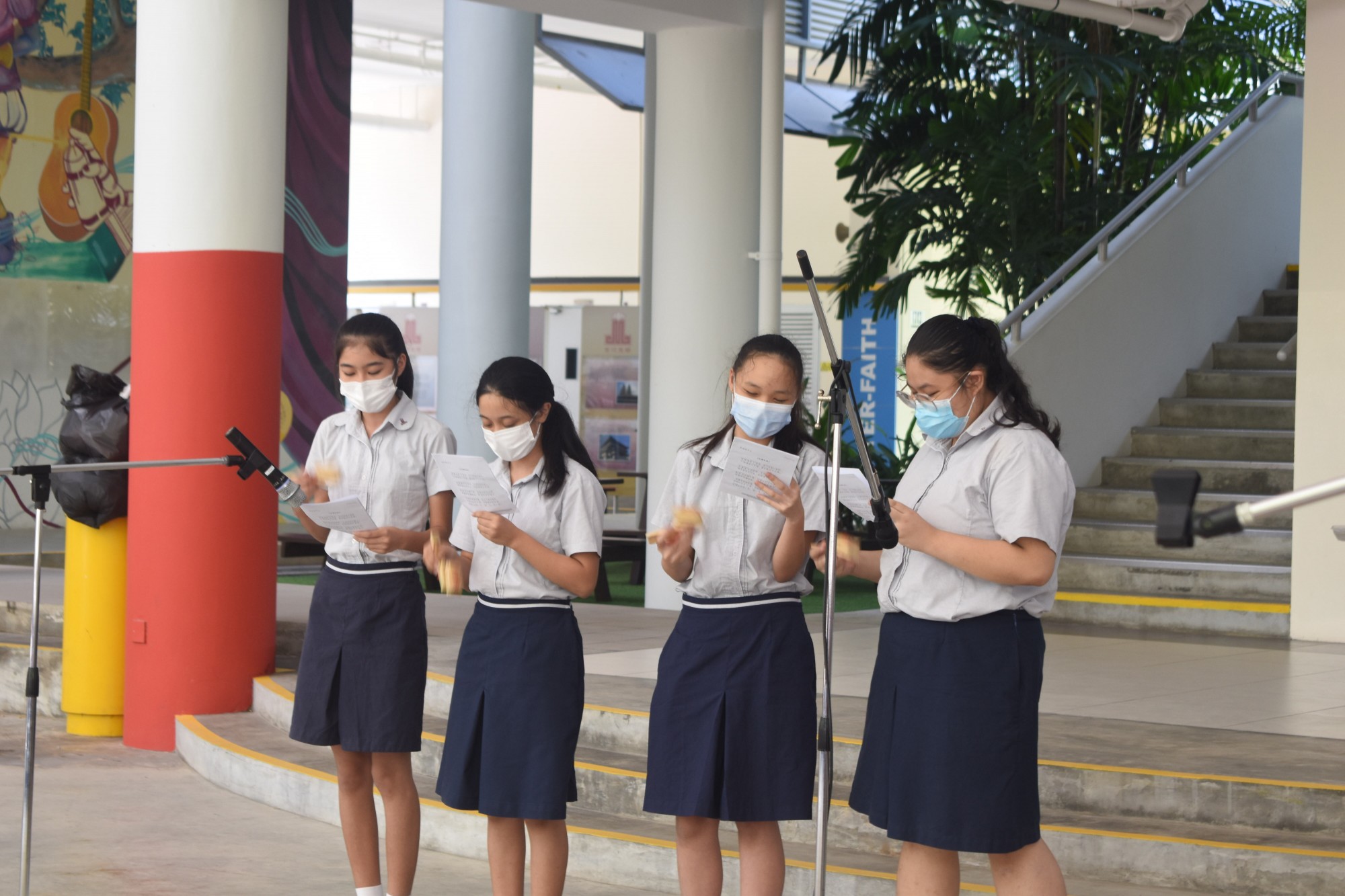 Four masked students performing with handbells and song sheets in a school event.