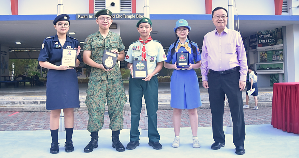Four uniformed youth cadets and an adult man pose outdoors holding award plaques near a National Cadet Corps sign.