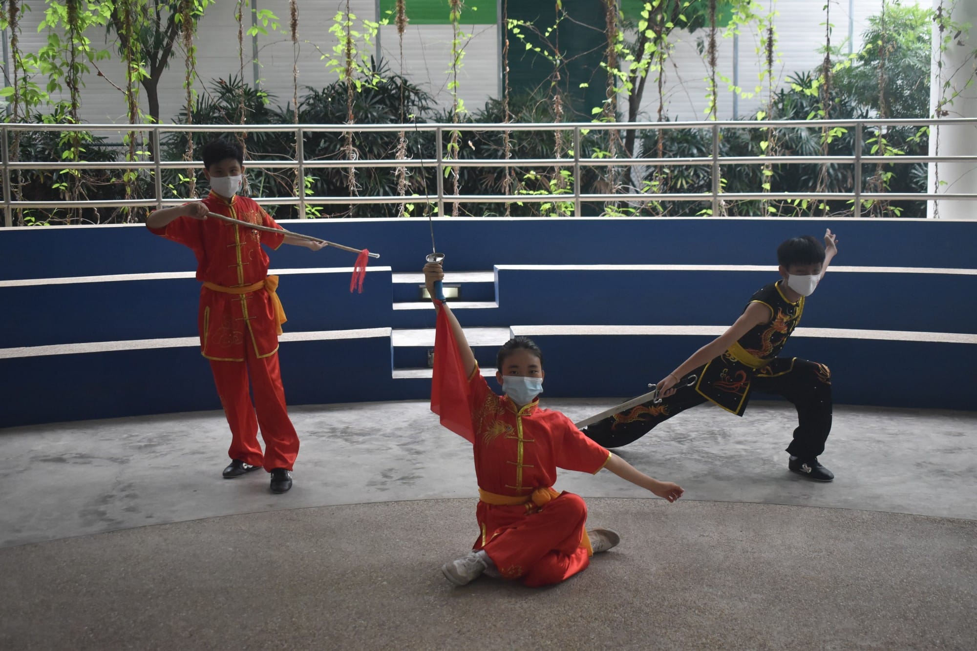 Three masked students in wushu uniforms performing martial arts poses with swords in a school courtyard.