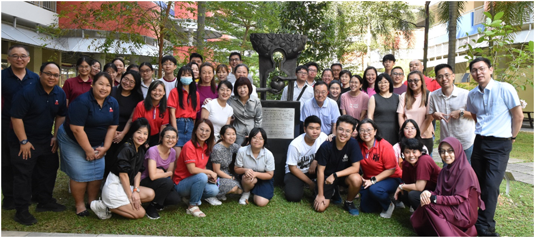 Group photo of students and staff posing outdoors around a sculpture in a school courtyard.