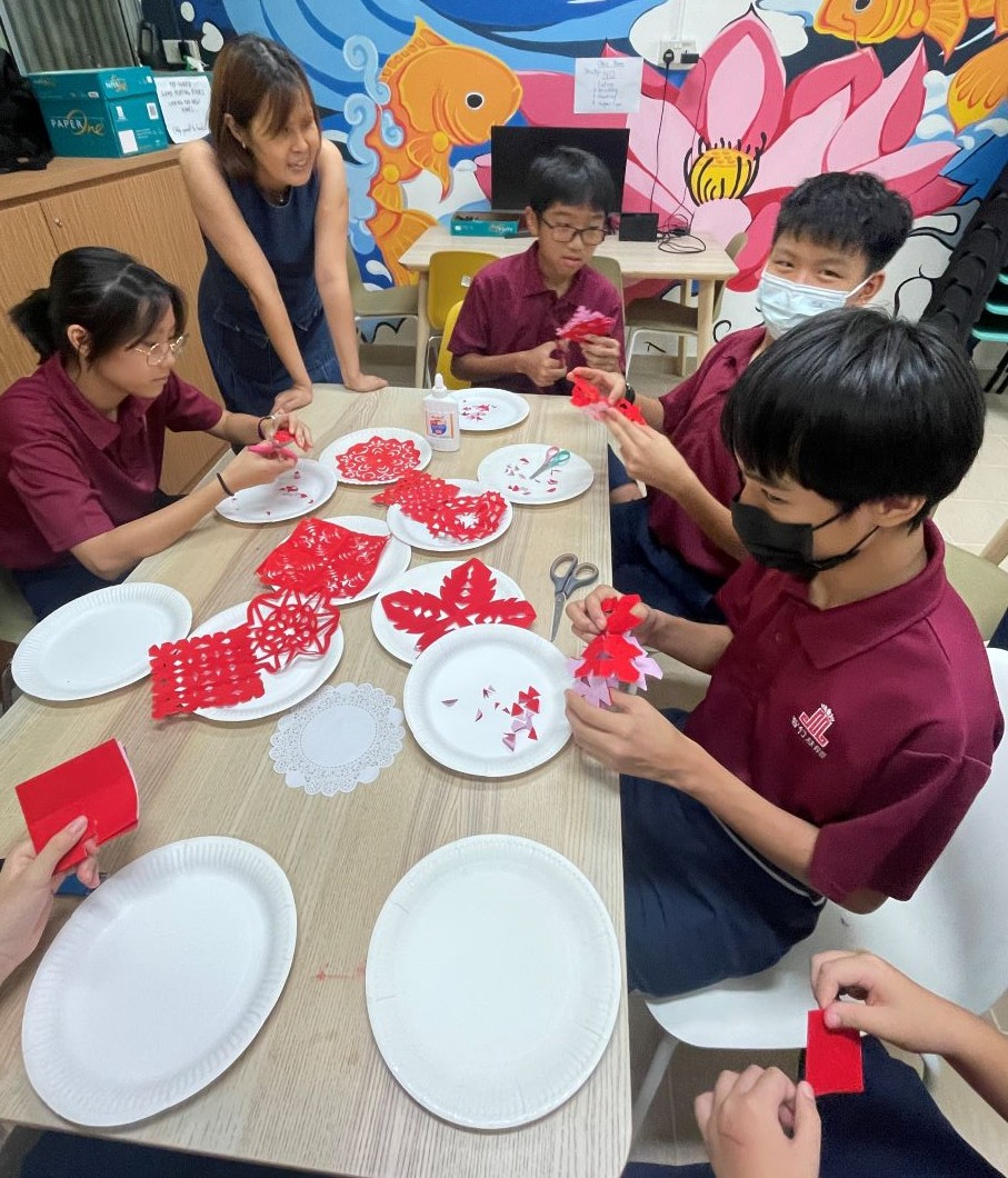 Children in a classroom working on a red paper craft project around a table, guided by a teacher