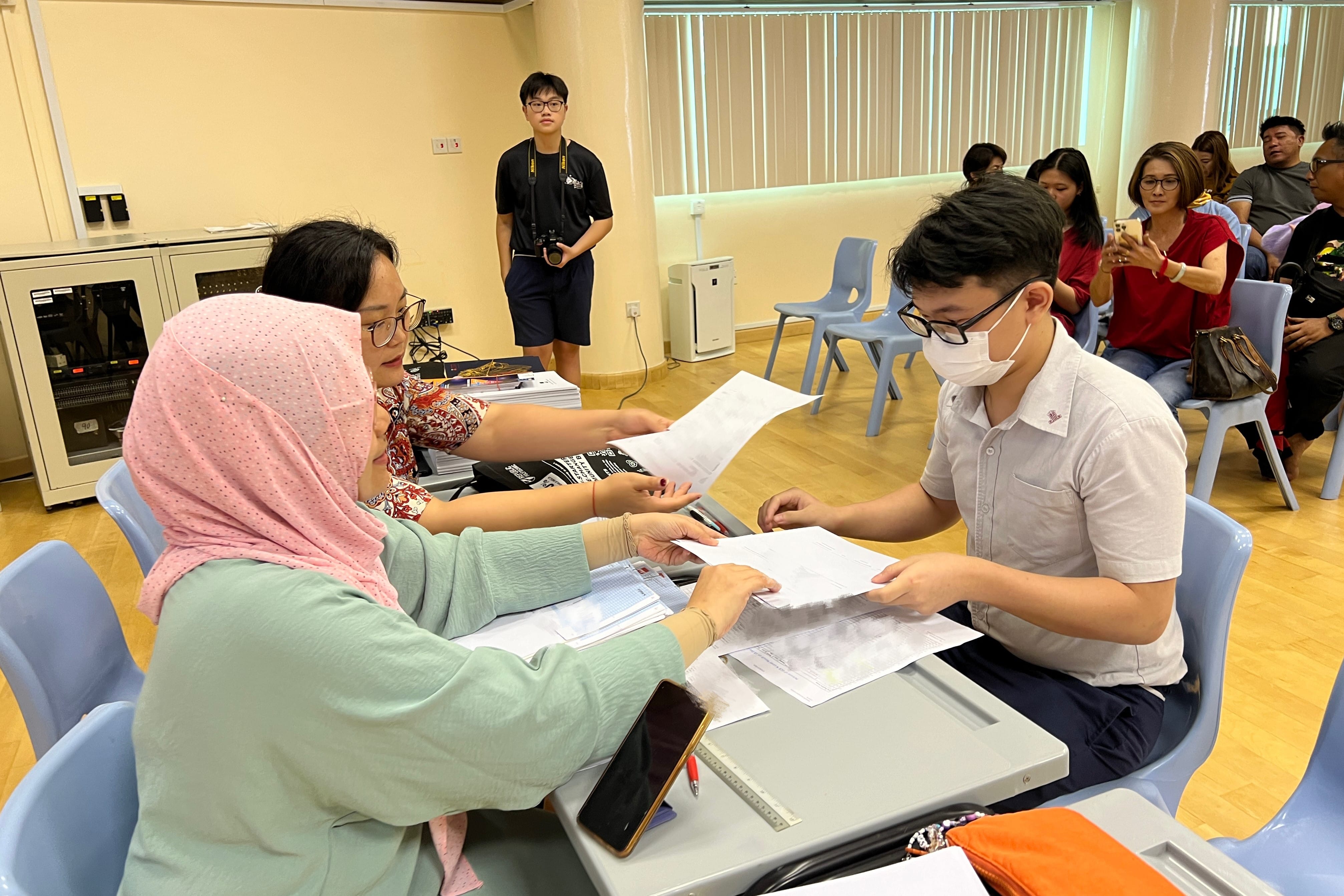 A student receives examination documents from staff as family members watch.