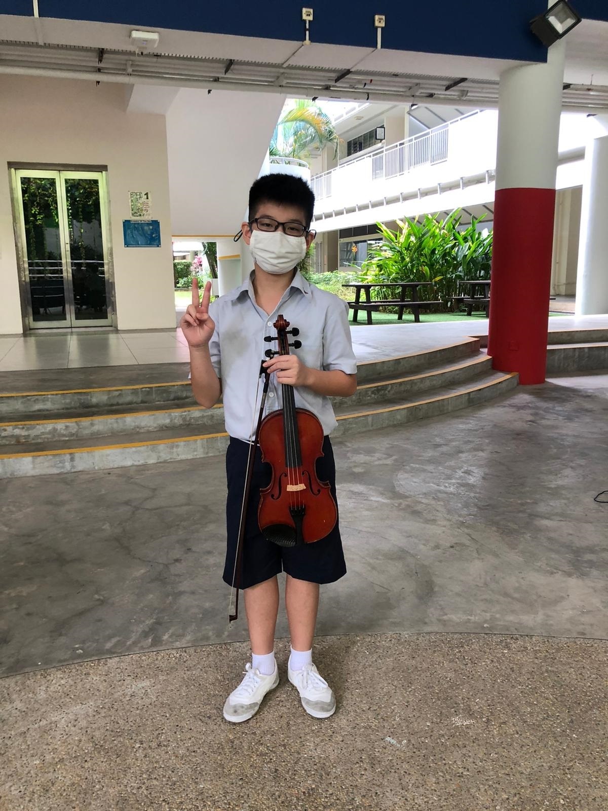 Student wearing a face mask holding a violin and making a peace sign in a school courtyard.