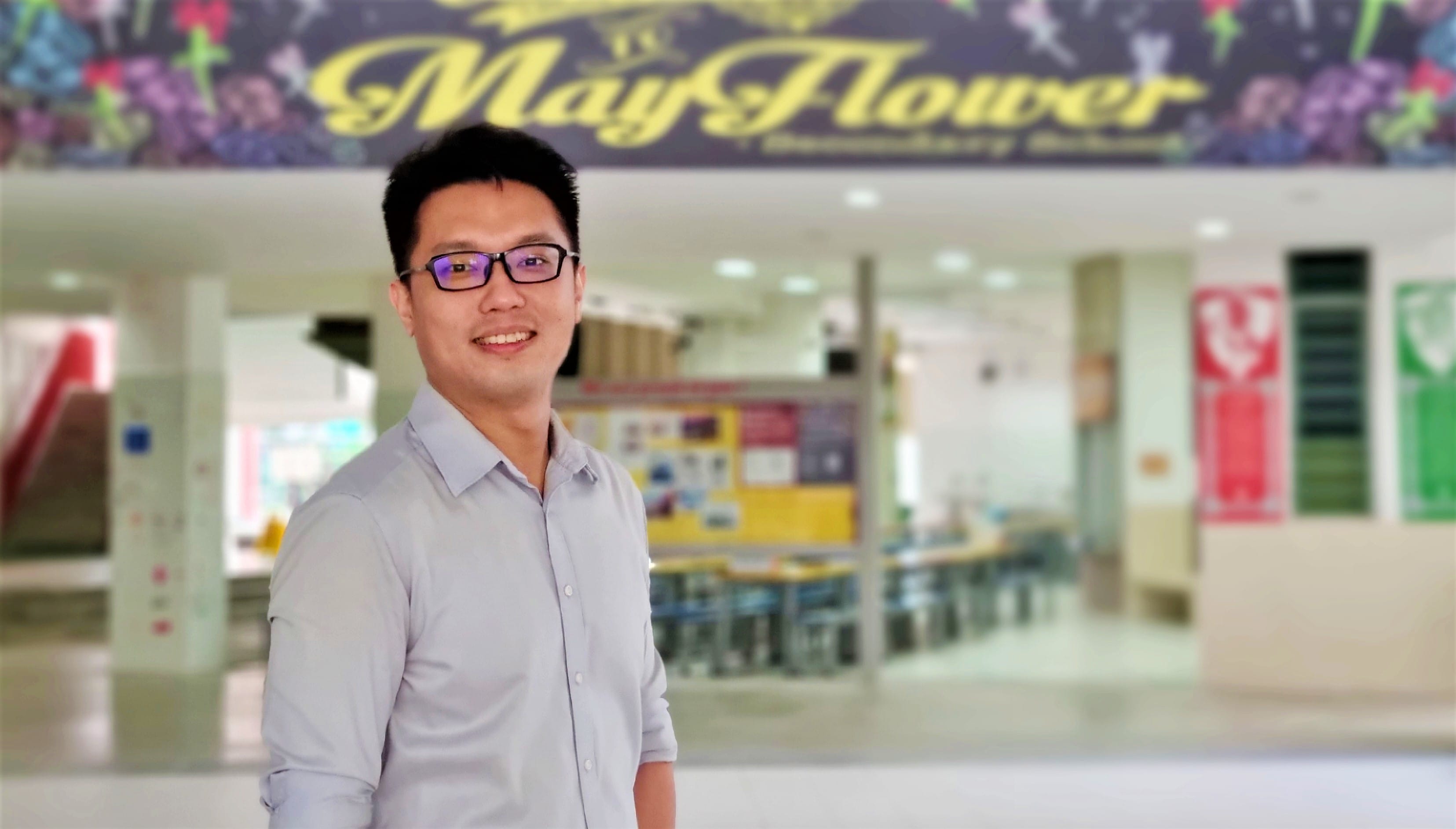 A smiling man wearing glasses and a grey shirt stands in front of a Mayflower Secondary School sign.