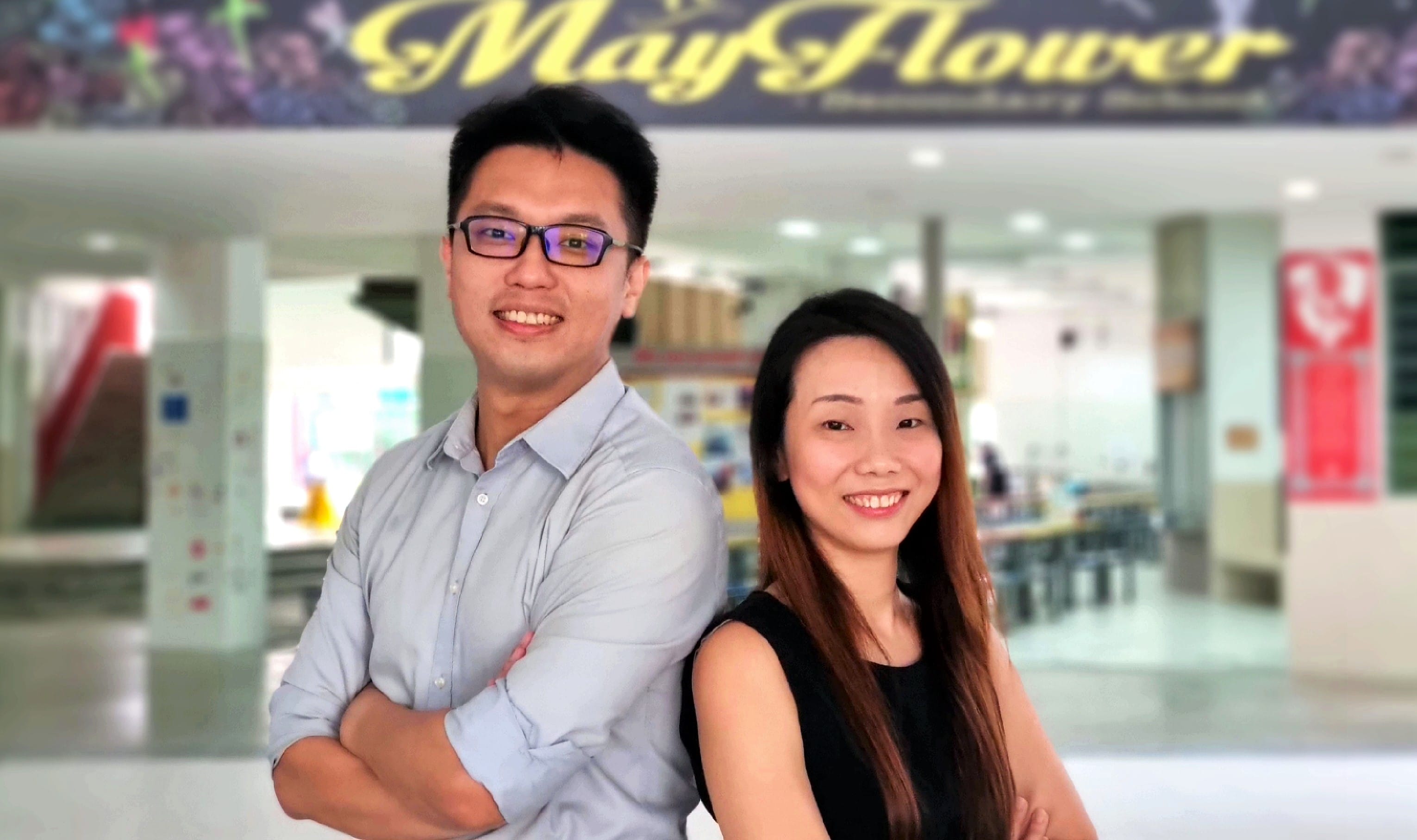 Two smiling people stand back-to-back in front of a Mayflower Secondary School entrance.