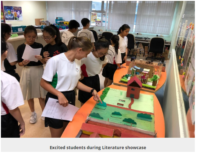 Students examine colorful handmade diorama models displayed on tables during a school literature showcase.