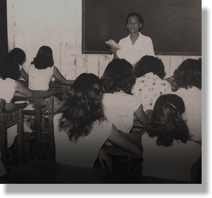 Black and white photo of a teacher holding papers standing before a chalkboard while students sit at desks.
