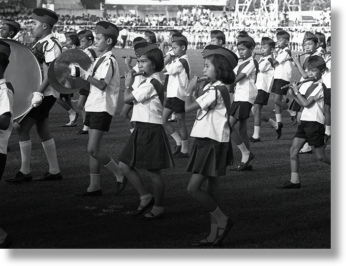 Children in uniforms and caps march in a parade, playing flutes and cymbals on a field before a crowd.