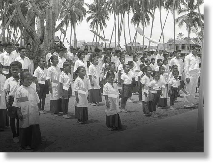 Black and white photo of children and adults in white uniforms holding small flags, standing in rows under palm trees.