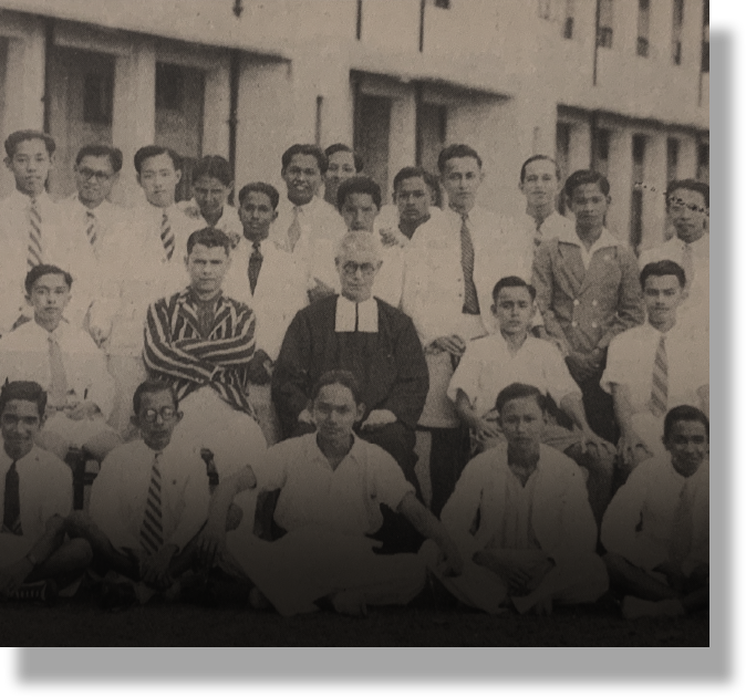 Vintage black and white group photo of young men posing with a clergy member in dark robes outdoors.