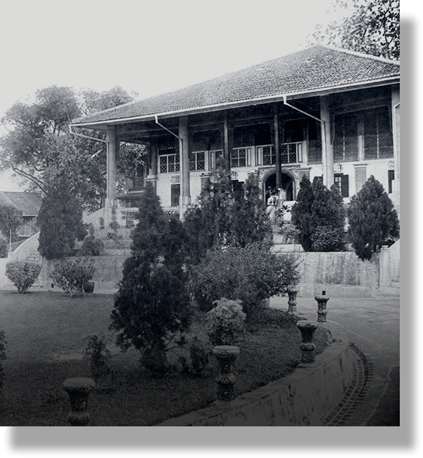 Black and white photo of a two-story colonial building with a tiled roof, surrounded by trimmed shrubs and a lawn.