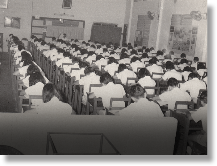 Black and white photo of a large hall filled with rows of students sitting at desks, appearing to take an exam.