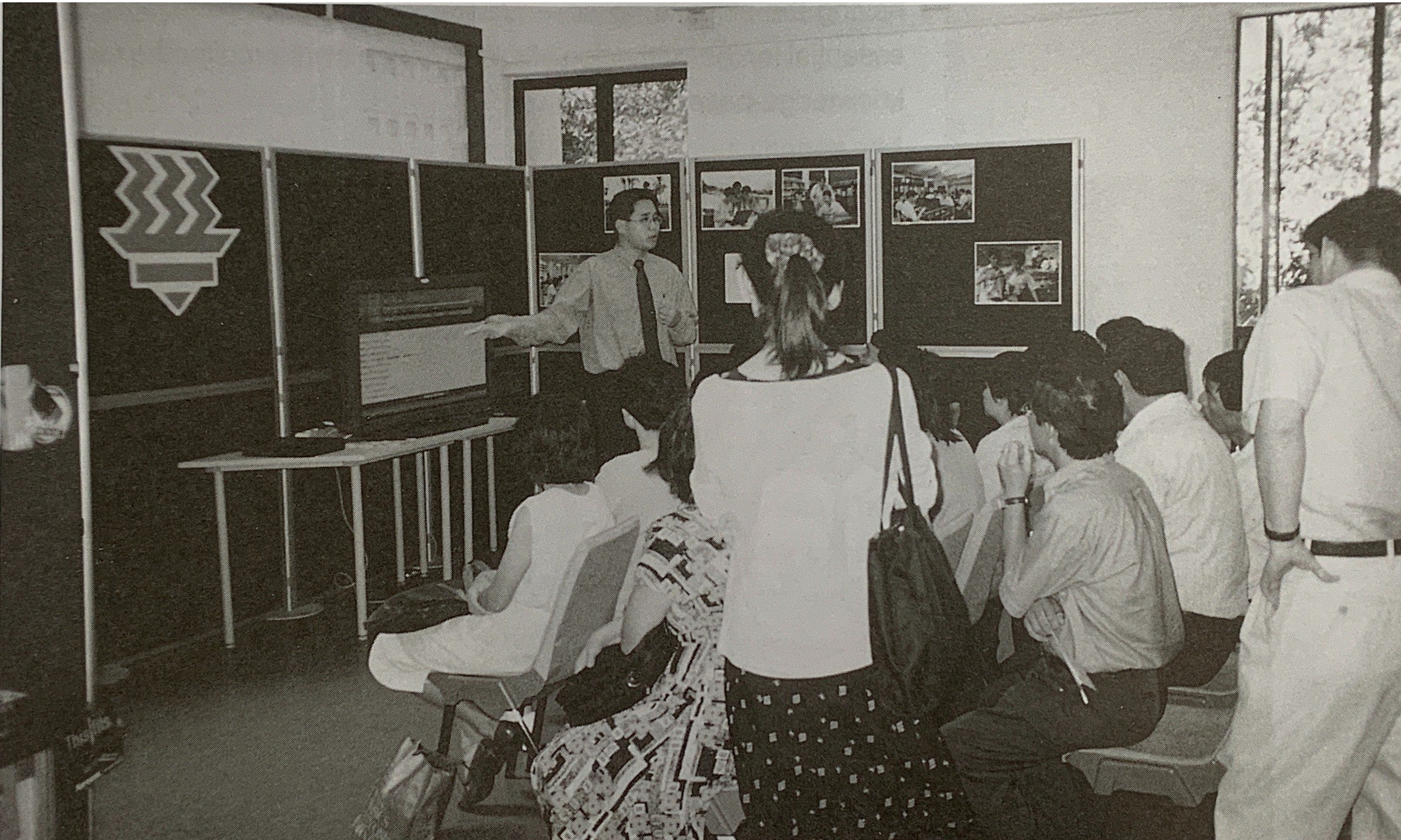Man presents at a screen and display boards as a seated audience watches in a room.