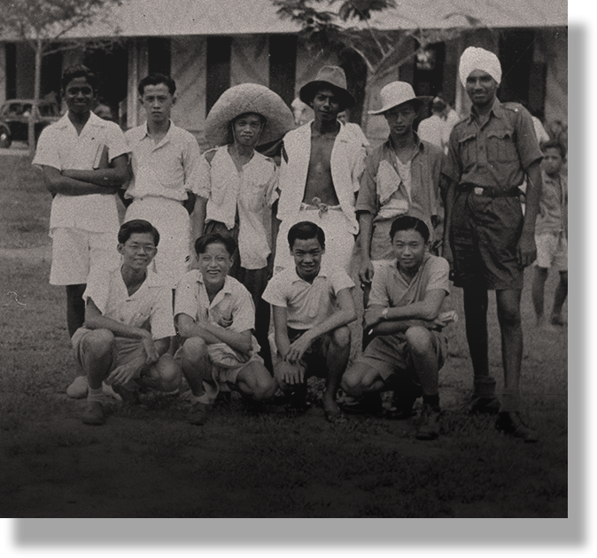 Black and white vintage photo of a group of about ten young men posing outdoors, some wearing hats, in front of a building.