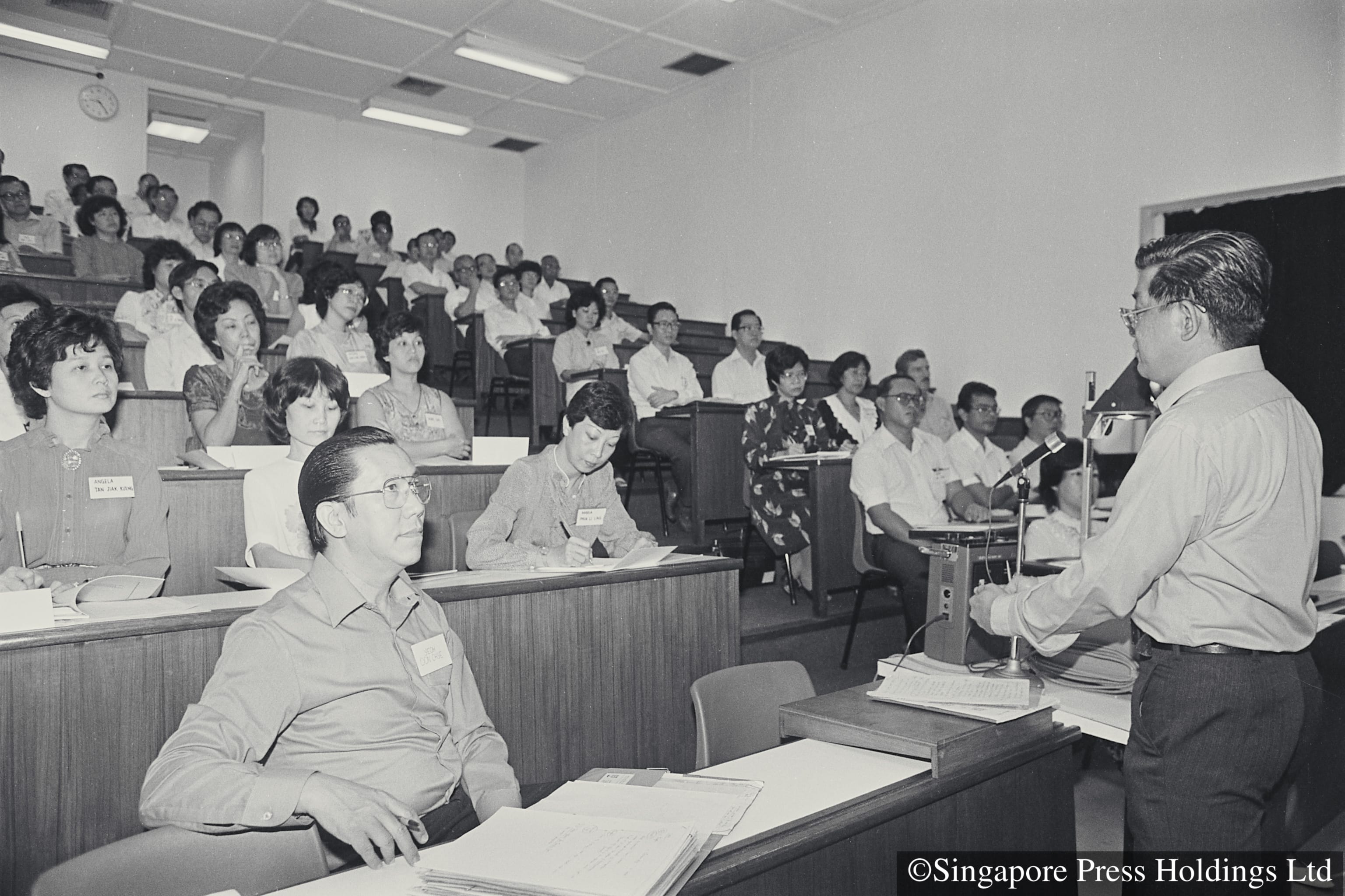 Black and white photo of a man speaking at a podium to a full lecture hall audience seated at tiered desks.