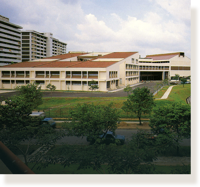 A wide low-rise building with red-brown roofs surrounded by green trees, a road with cars, and tall apartment blocks nearby.