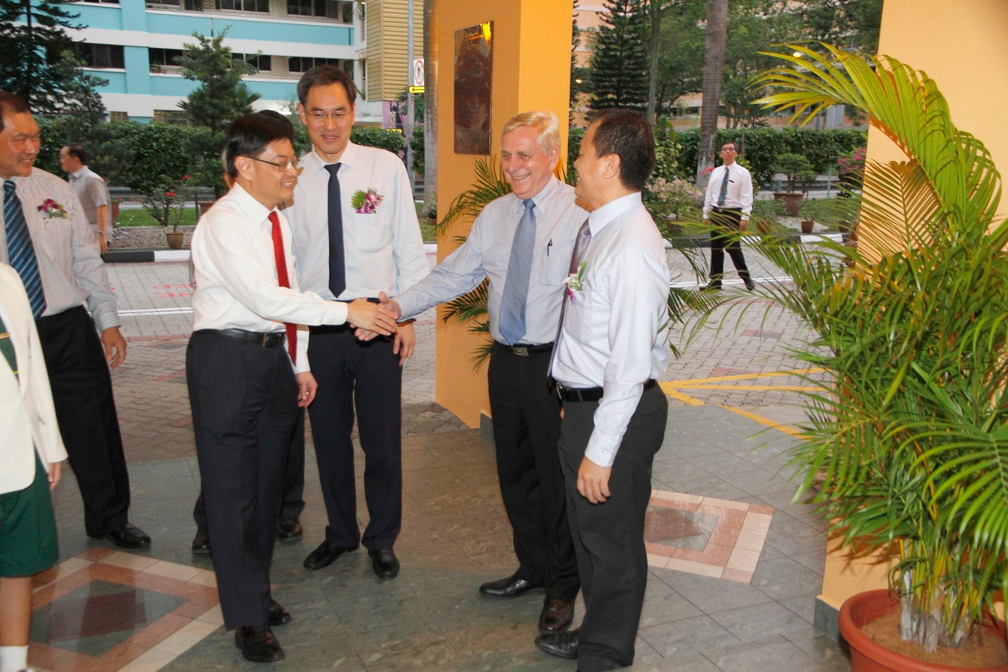 Four men in dress shirts and ties shake hands outdoors near a yellow building, with trees and a palm plant nearby.