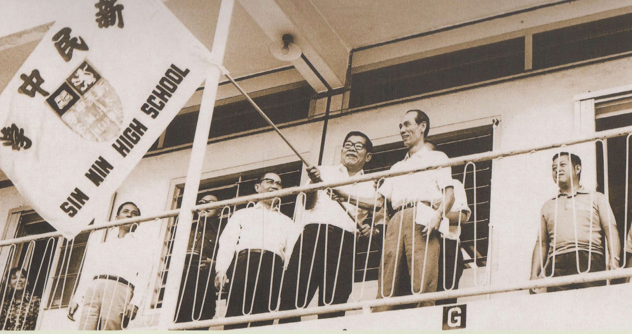 Sepia photo of men on a building balcony, one holding a flag reading Sin Min High School with Chinese characters.
