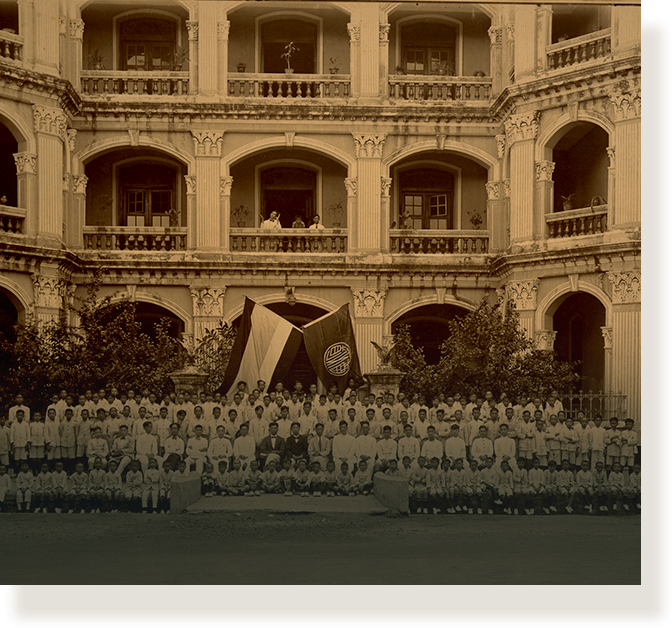 Sepia-toned vintage photo of a large group of people posed in front of a grand multi-story colonial building.
