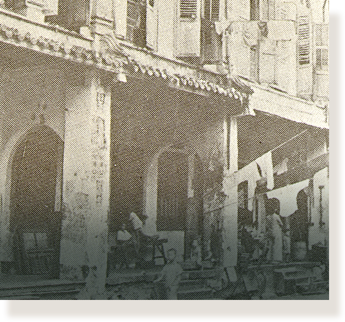 Old sepia photo of a colonial-style building with arched columns and laundry hanging outside, people walking below.