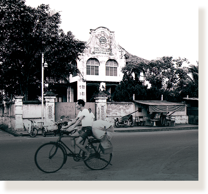 Black and white photo of a person cycling past an old colonial school building dated 1912, with trees nearby.