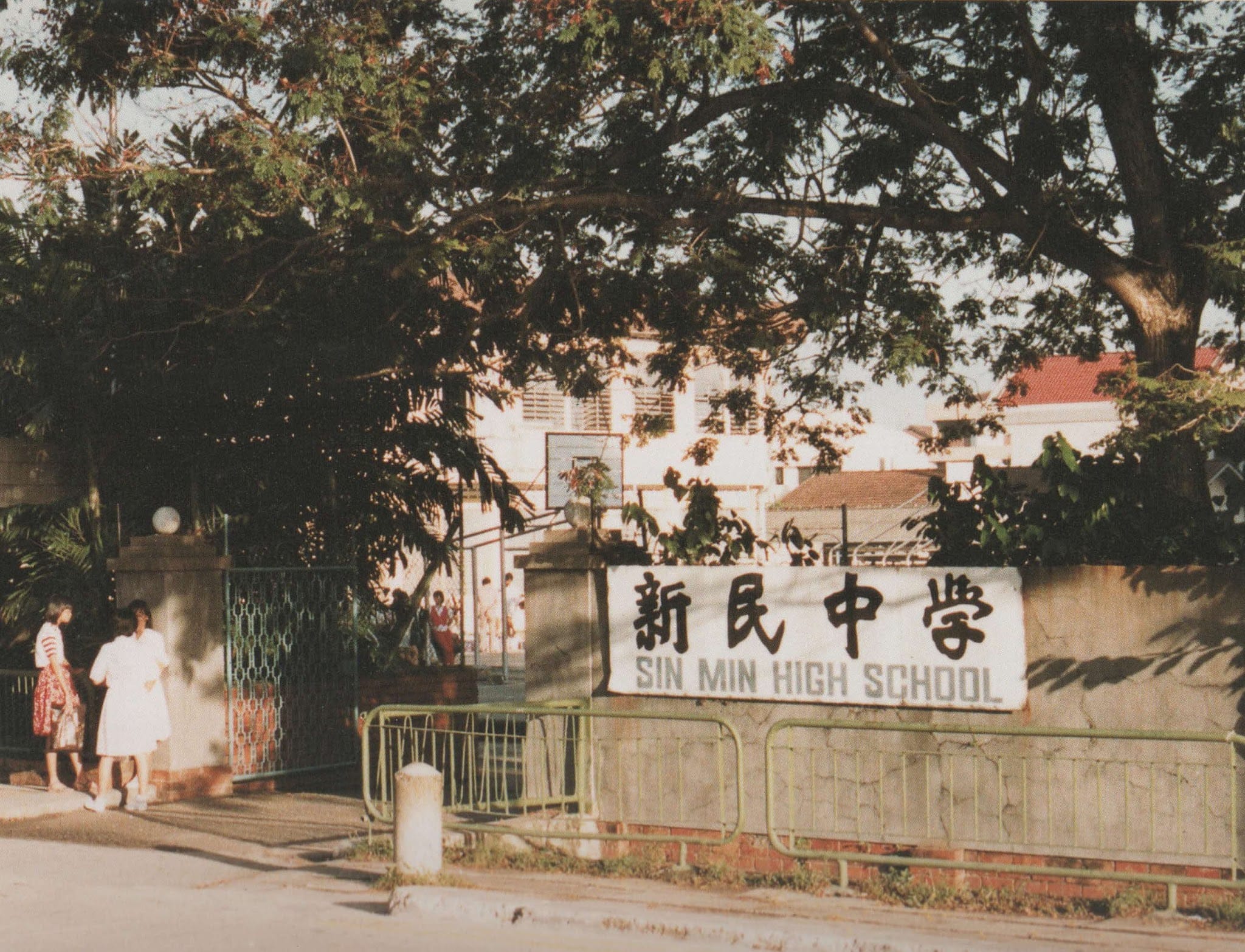 Entrance of Sin Min High School with Chinese characters on a wall sign, two people near a gate, and large trees overhead.