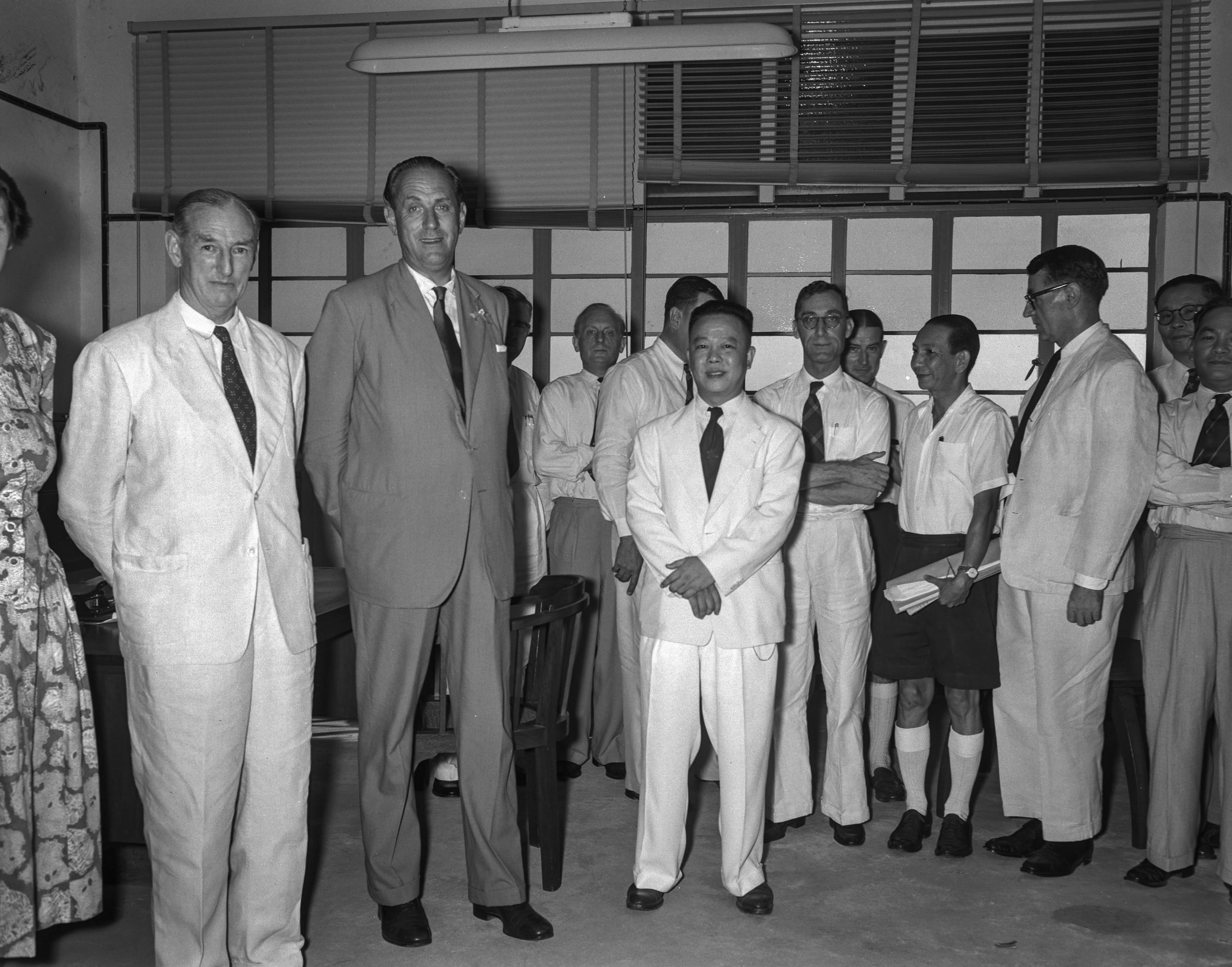 Group of men in suits and light jackets stand in an office room, posed for a black-and-white photo.