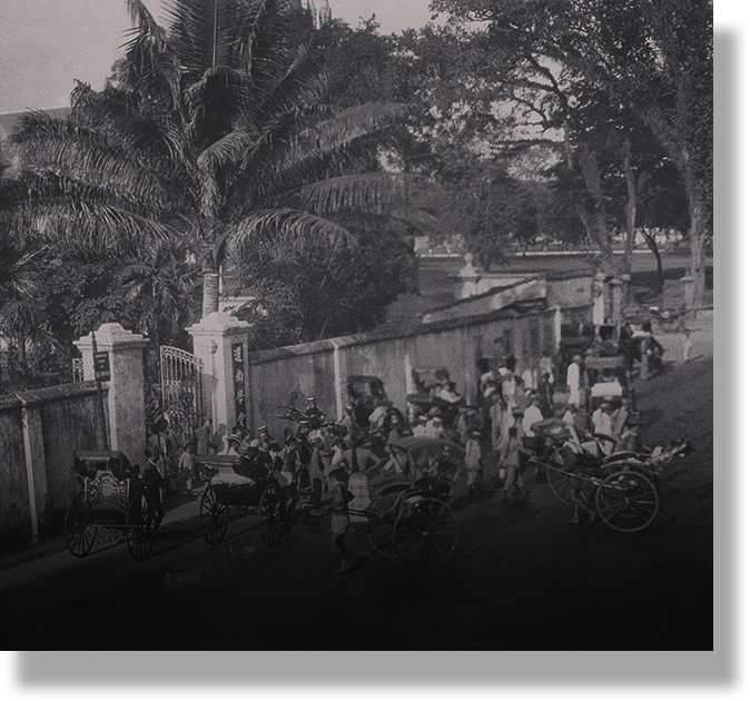 Old black and white photo of a crowded street with rickshaws, people, a gated wall, and palm trees.