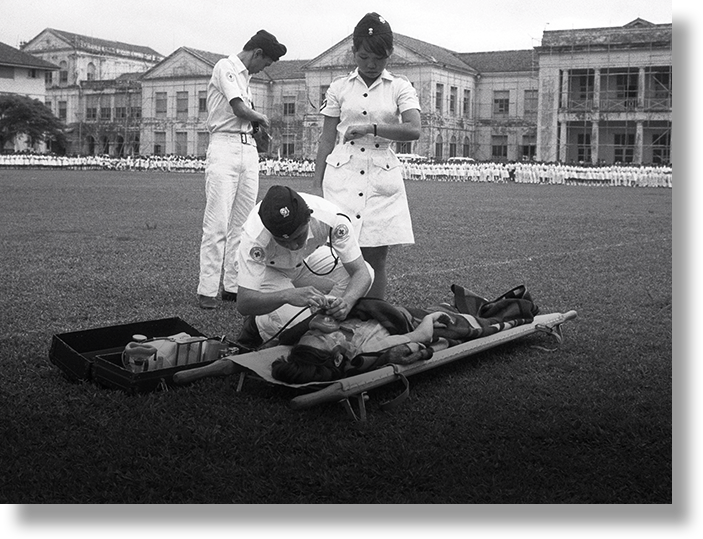 Three medical personnel in uniform attend to a person lying on a stretcher on a field, with a crowd in the background.