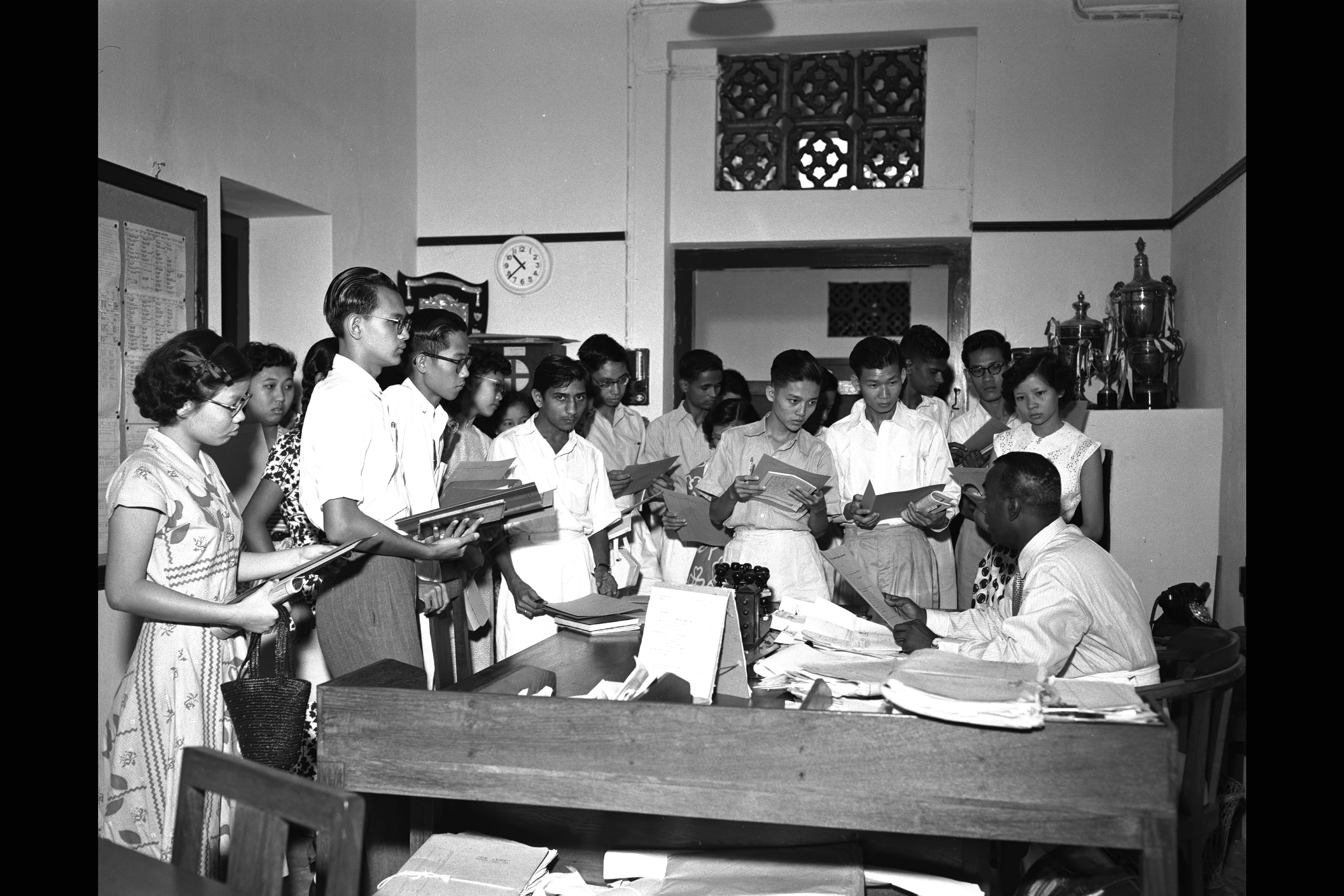 Black and white photo of a group of young students holding papers, standing before a man seated at a desk in an office.