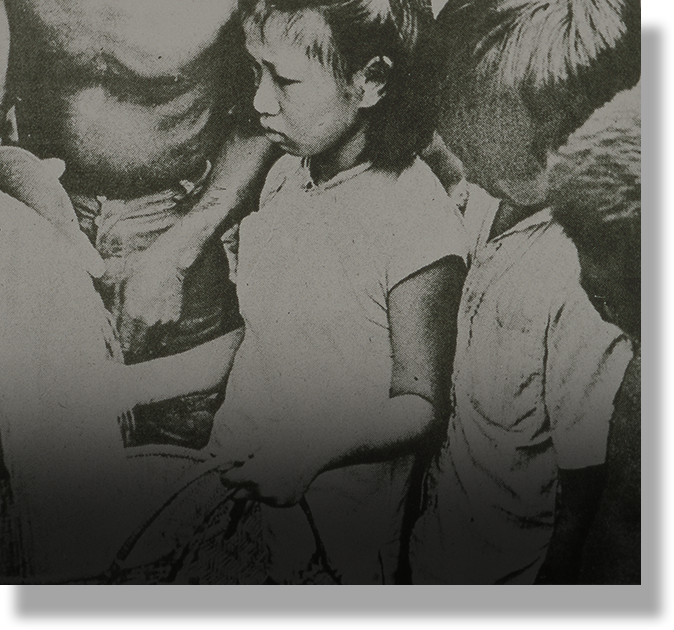Black and white photo of two young children standing close together, with an adult partially visible behind them.