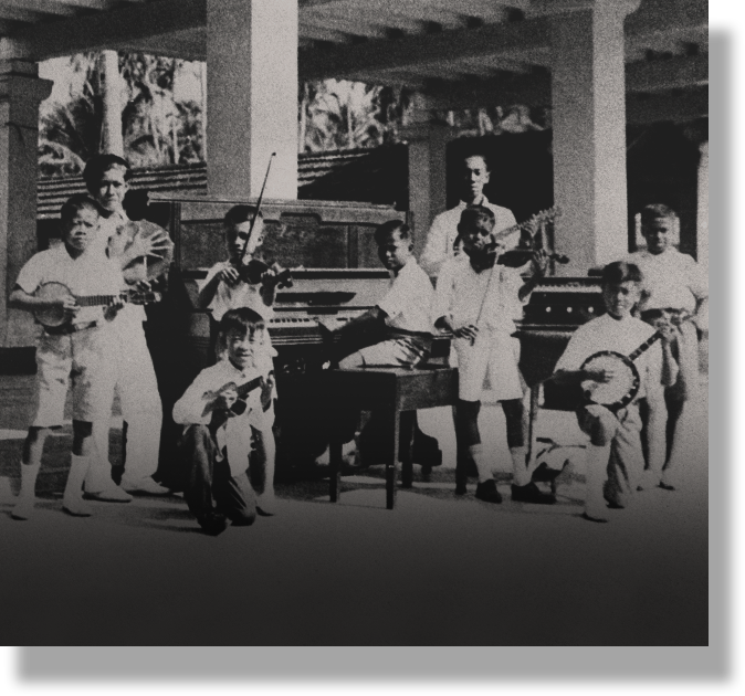 Black and white vintage photo of a group of young musicians holding banjos, violins, and a piano outdoors under a covered area.
