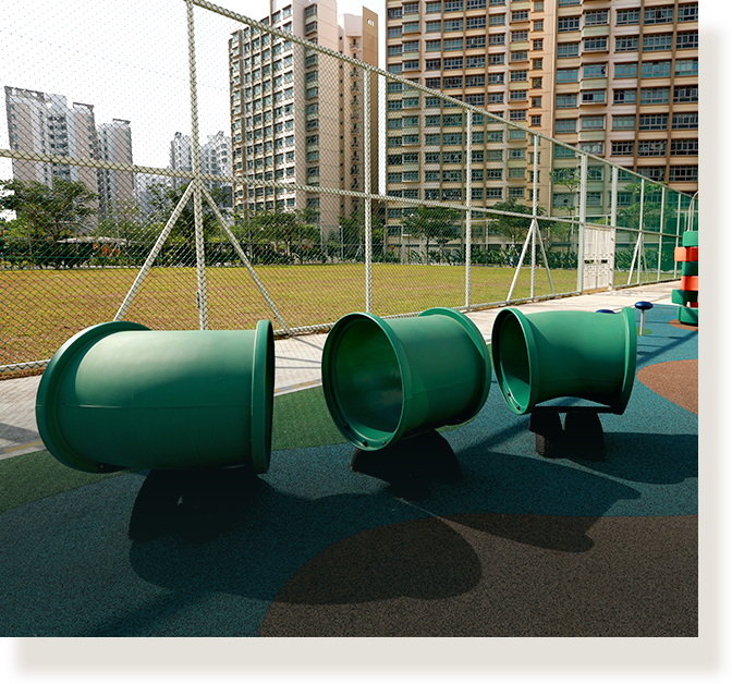 Green tunnel crawl tubes on a rubber playground surface, with a chain-link fence and tall apartment buildings behind.