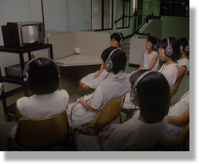Several children wearing headphones sit in chairs watching a small CRT television in a room.
