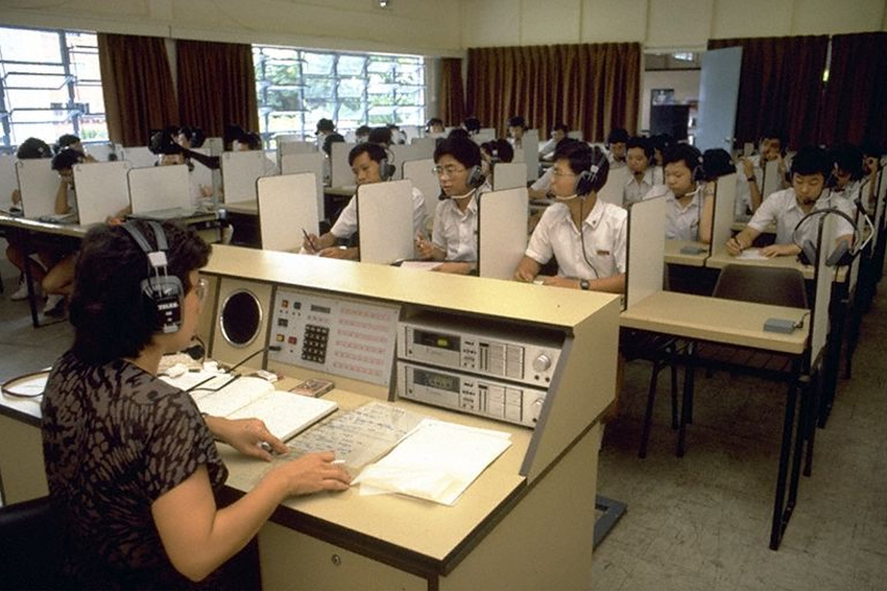 A teacher at a control desk and many students wearing headphones sit at partitioned desks in a language lab.