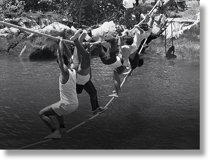 Several people hang from a rope bridge crossing over a river, with rocky shores and trees in the background.