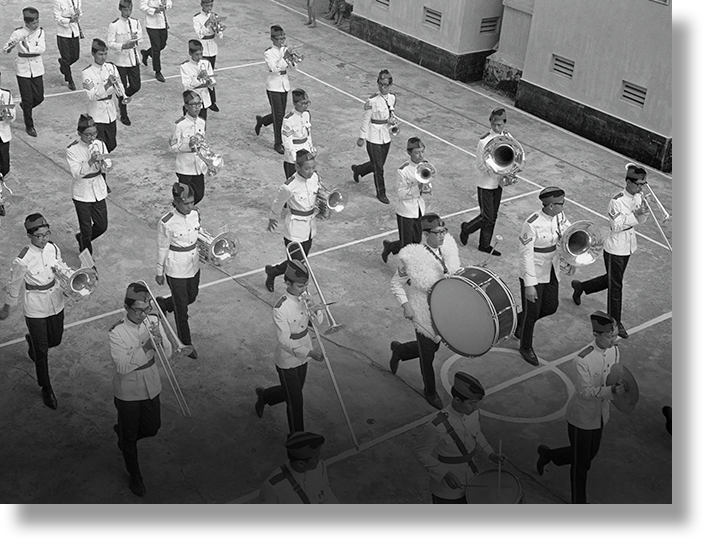 Black and white photo of a youth marching band in white uniforms playing instruments on an outdoor court.