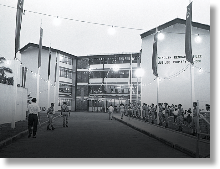 Black and white photo of Jubilee Primary School exterior at dusk, with uniformed personnel and string lights.