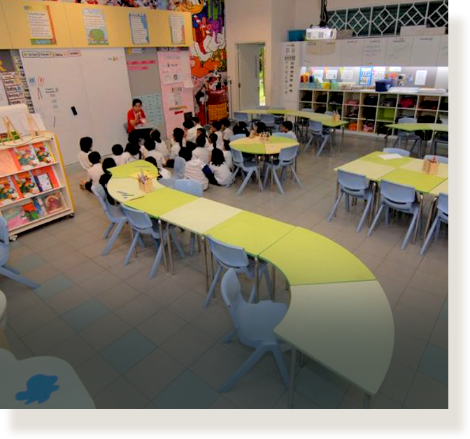 Young children in white uniforms sit on the floor facing a teacher in a bright, colorful classroom.