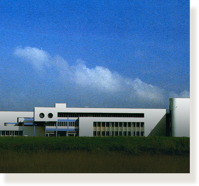 A low, wide white modern building with circular windows set against a blue sky with clouds, surrounded by grass.