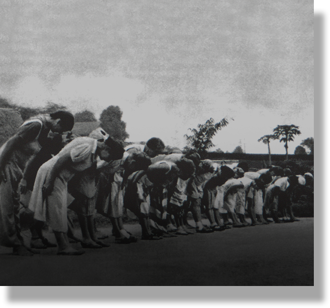 Black and white photo of a long line of people bowing forward outdoors, with trees visible in the background.