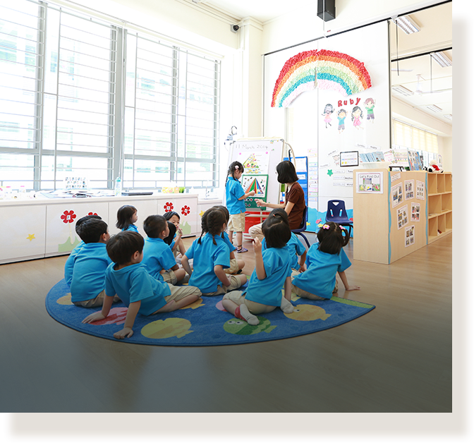 Young children in blue uniforms sit on a rug facing a teacher at a whiteboard in a bright classroom.